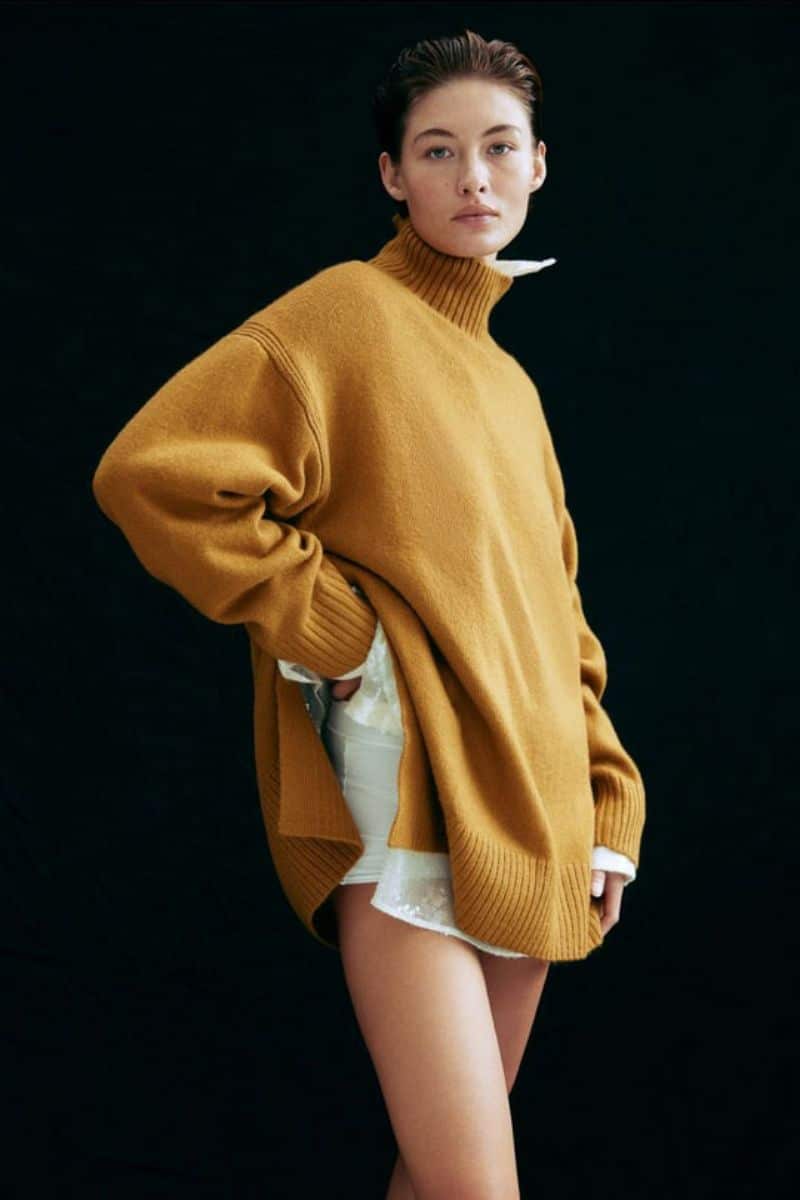 Model wears yellow oversized turtleneck in front of black background. 