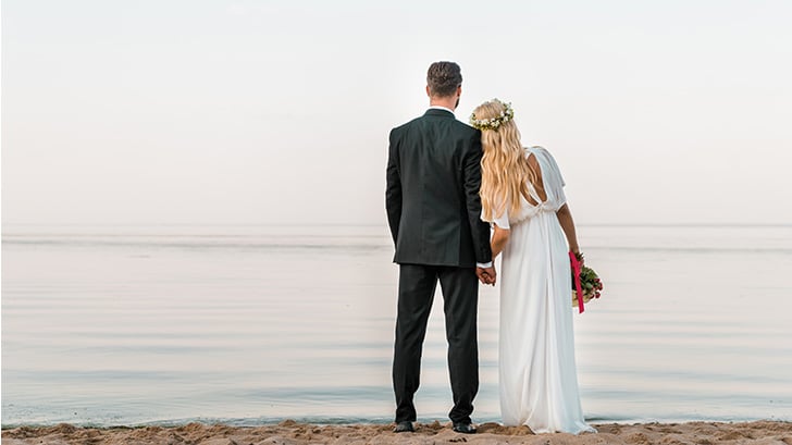 The best money saving wedding tips, according to experts 1 View from behind of bride and groom standing on the beach.