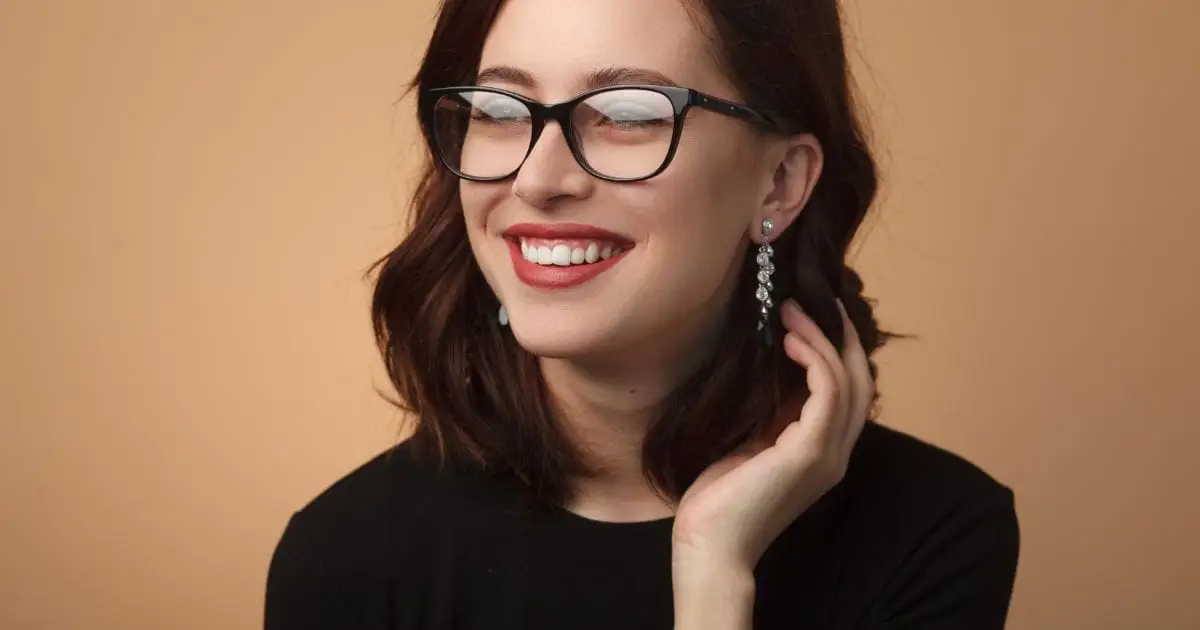 Smiling woman wears eyeglasses in front of tan background.
